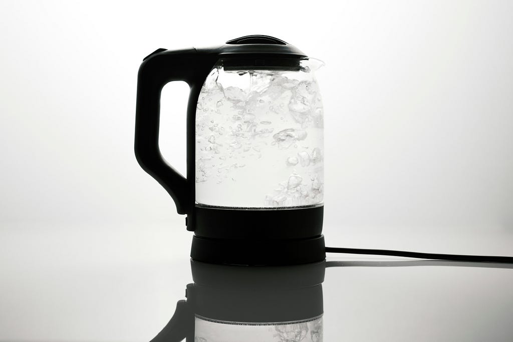 A transparent kettle with black plastic handle, lid and base, is filled with boiling water.