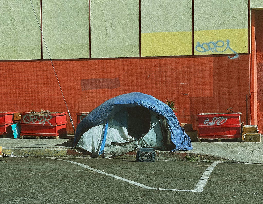 A black-asphalt parking lot abuts a sidewalk running the length of a painted red-brick. big-box building with windows papered over from the inside. On the sidewalk sits a blue dome tent, its door-flap open.