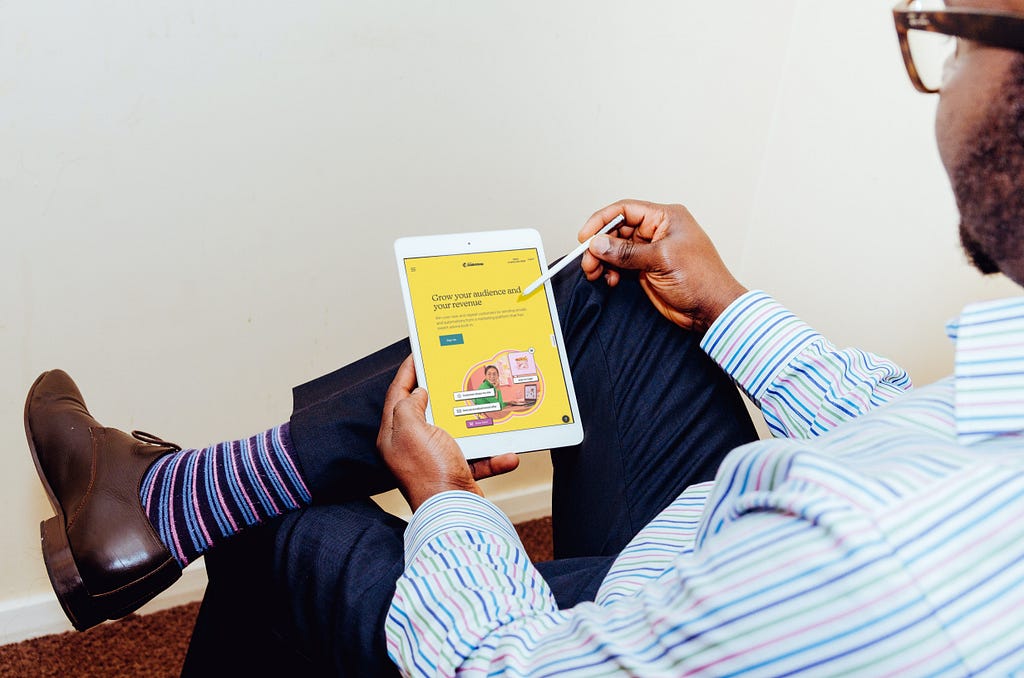 Man with a blue striped shirt sits with one-leg over the other while he views a tablet screen.