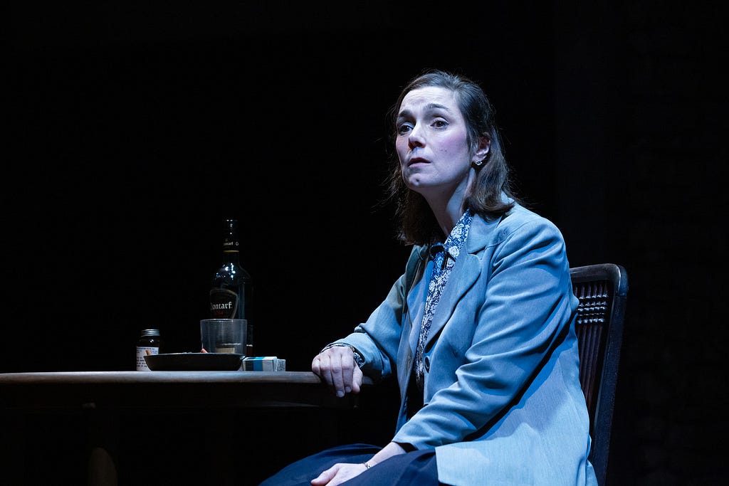 A woman in a floral blouse and gray suit jacket sits at a table with an ashtray and a bottle of whiskey in blue light.