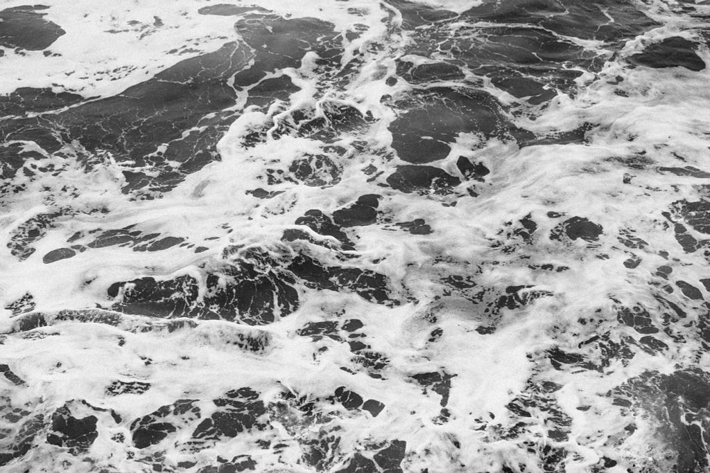 A black and white shot of the sea, rippling and creating the characteristic foam.