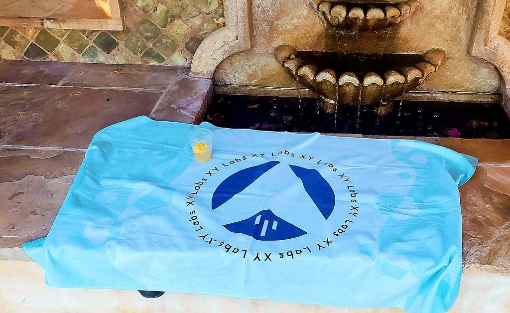 Blue beach towel with XYO World graphics and the XY Labs logo and name laid out on a stonework bench with an embedded Spanish-style fountain. A yellow cocktail in a plastic cup sits on the towel.
