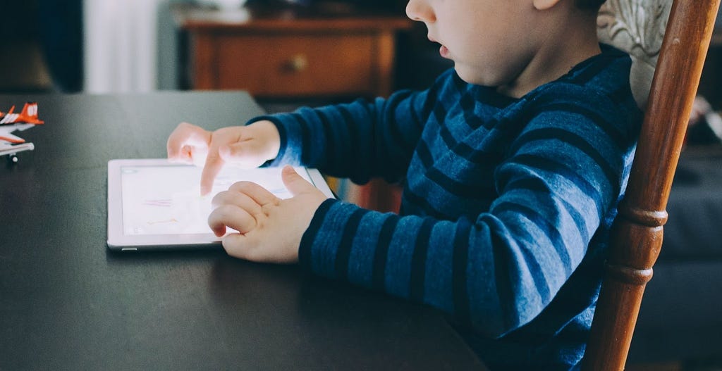 A child sits at a kitchen table, tapping on the glowing screen of an iPad