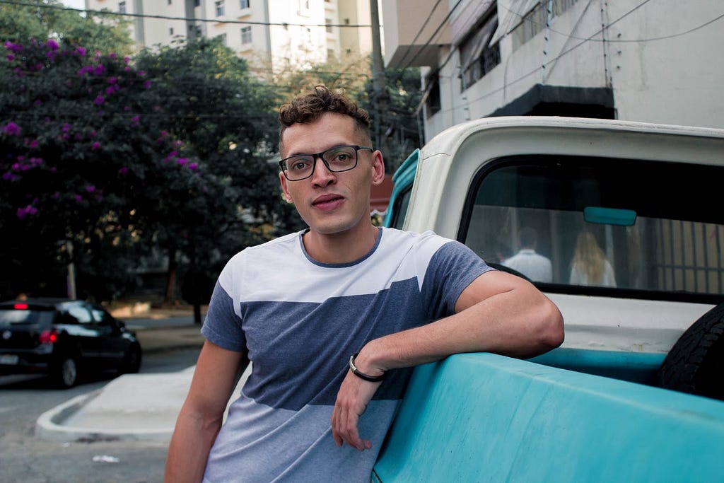 Imagem de perfil do Matheus. Ele usa camiseta cinza e branca, óculos e está encostado em um carro azul. Ao fundo uma rua.