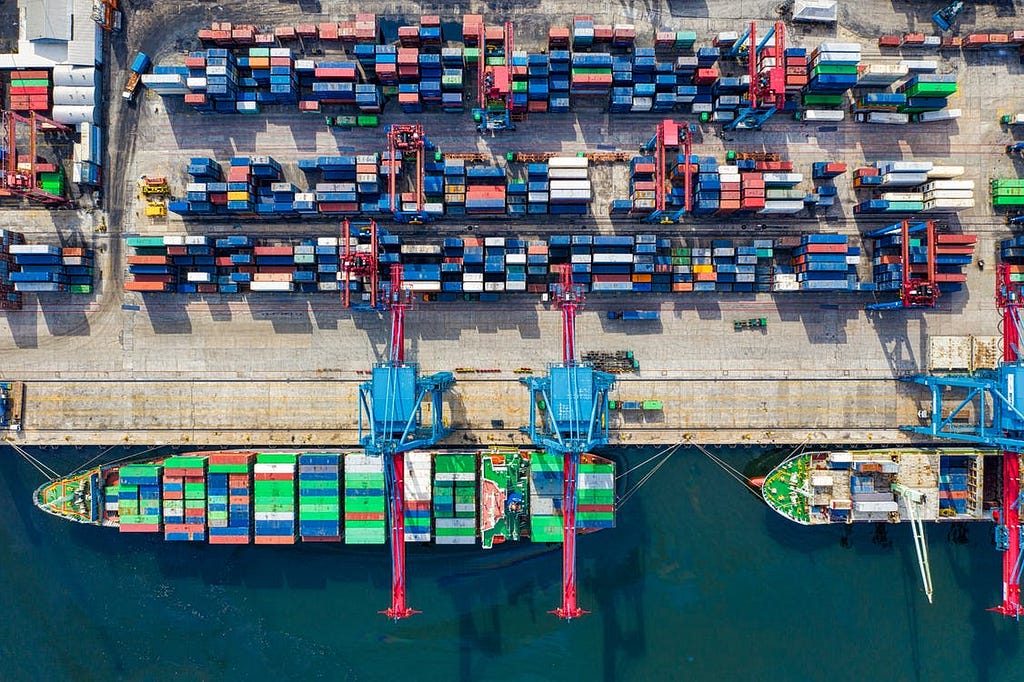 An aerial view captures a shipyard with multiple containers. A liner is shown at full capacity docked at the shipyard.