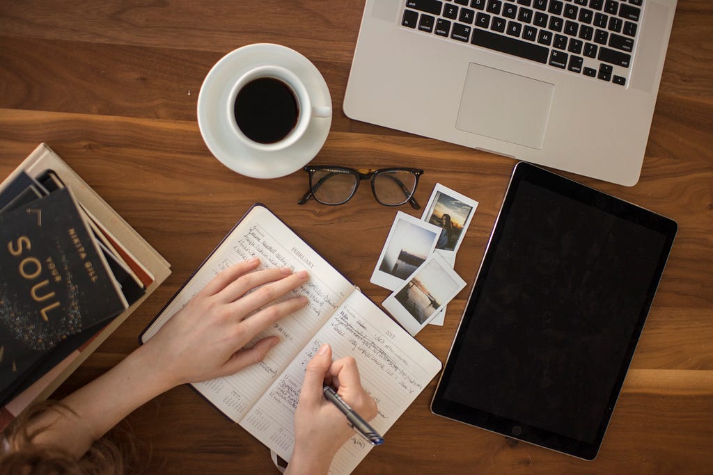 A visual of a person writing on a notebook with coffee, laptop, polaroid pictures, and books.