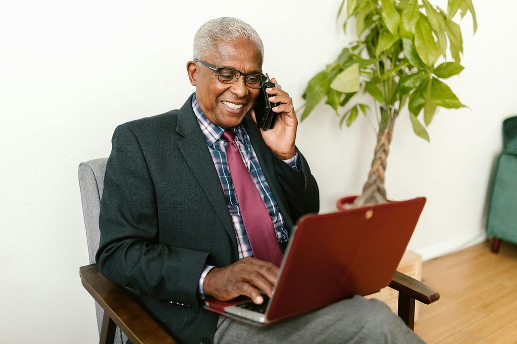 A male senior citizen sitting on a chair, looking at his laptop while talking on his smart phone