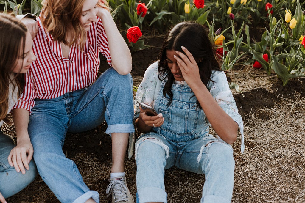 Three girls sitting on soil socializing and smiling while the girl on the far right scrolls through her cellphone