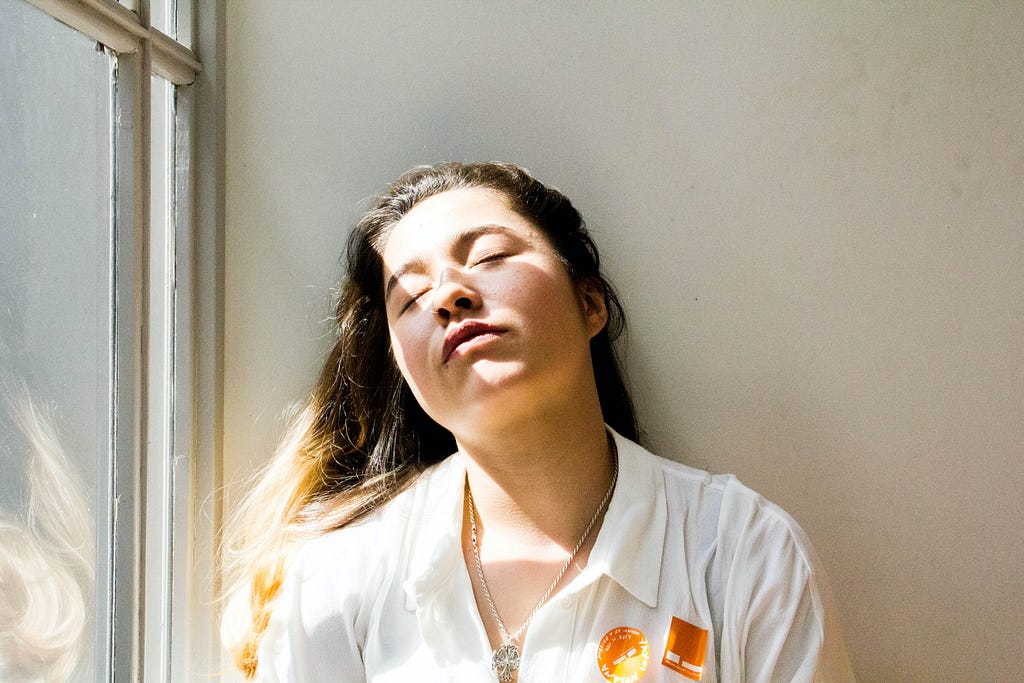 Dark-haired woman with a white blouse next to a window has her eyes closed and head against the wall has a facial expression that might say, “Oh,  I’m so very tired, it’s a sunny day and my eyes will not stay open.