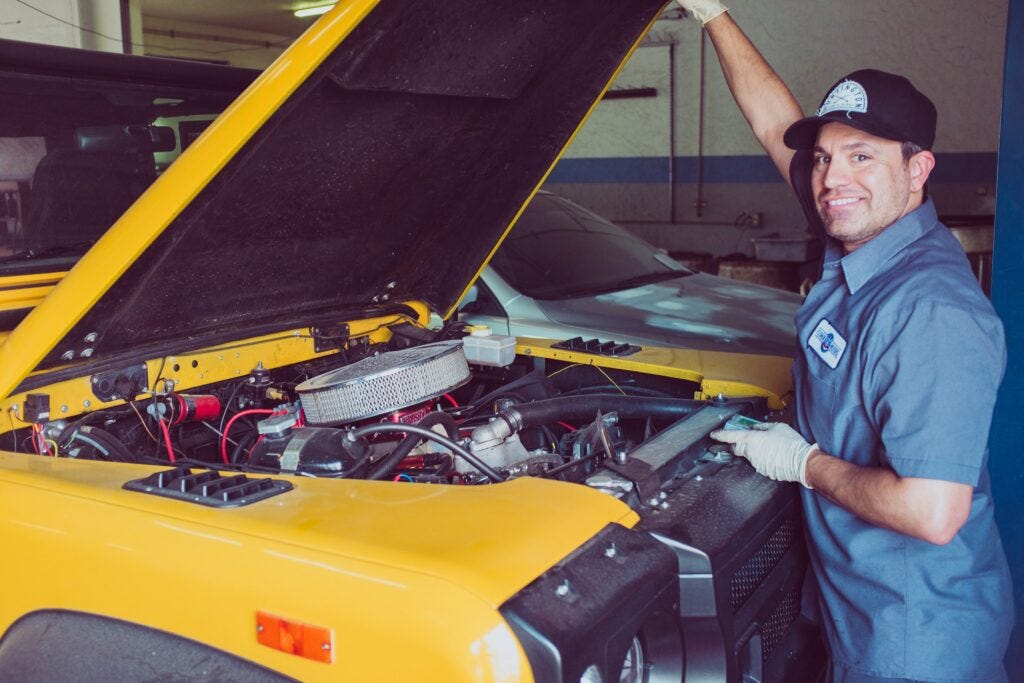 Un mecánico sujeta sonriente el capó de un Land Rover Defender amarillo. El mecánico viste camisa vaquera azul, gorra negra, y guantes de faena blancos.
