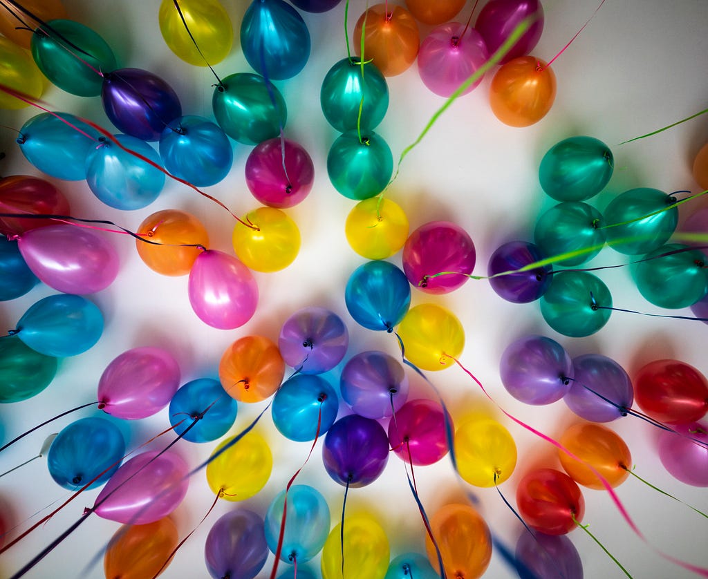Dozens of shiny balloons in a wide range of colors push against a white ceiling, photographed from below, their shiny ribbon strings dangling towards the camera and radiating from the image center