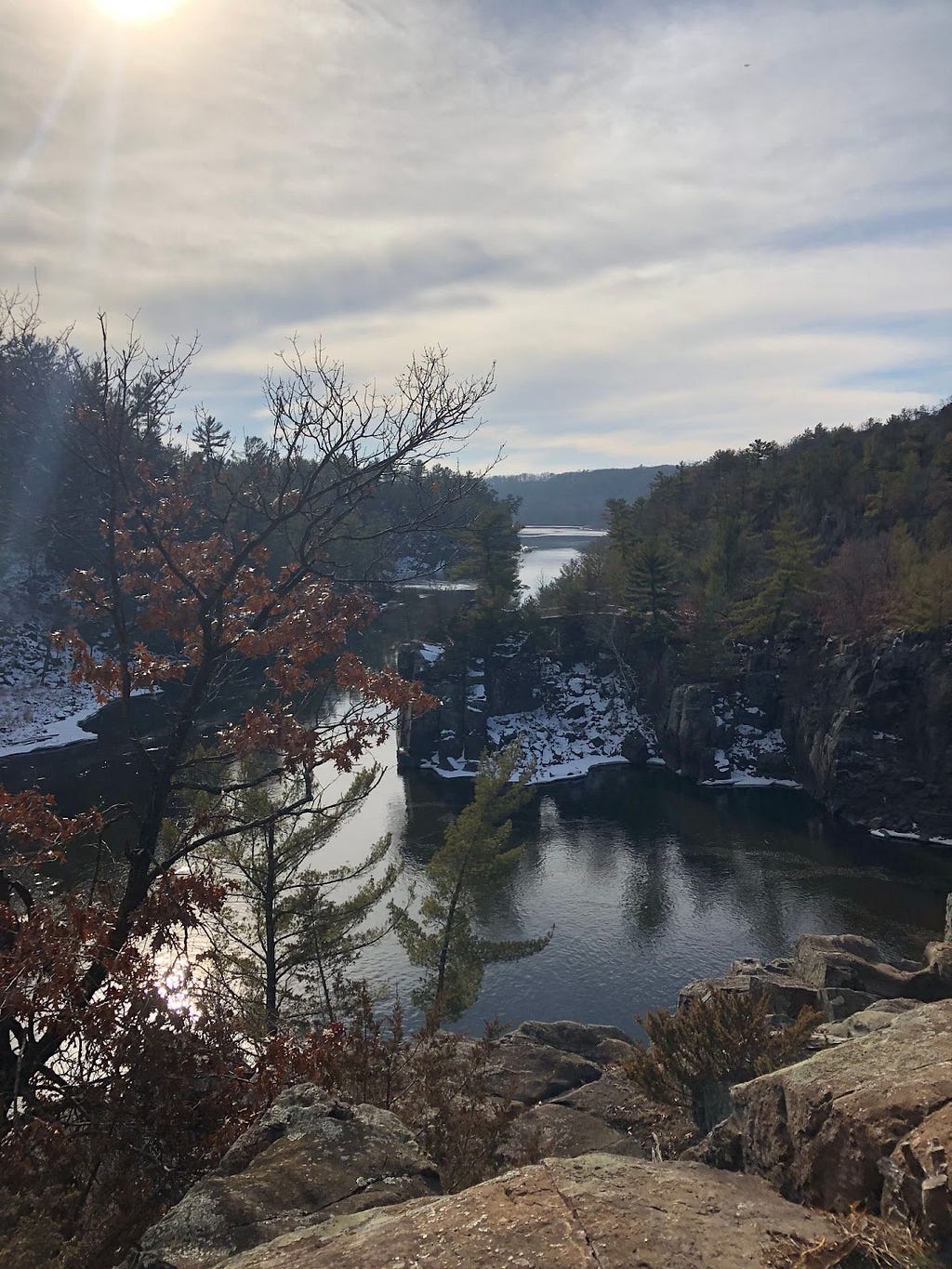 Scenic view of the Dalles in Taylor Falls: a river flowing through a rocky gorge with trees lining the banks, snow covering the lower edges, under a cloudy sky.