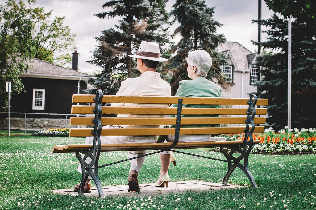 A man of retirement age wearing a Panama hat sits on a slat bench with his female companion peering across a green area covered in green grass and clover within view of homes and a colorful flower bed.