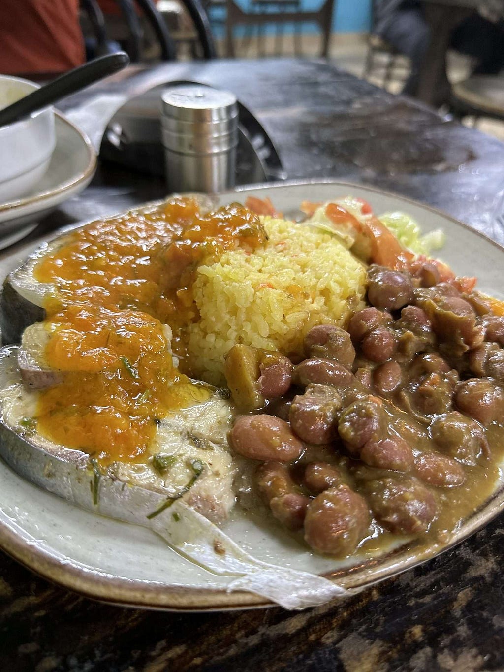 A white plate with rice, cooked beans, and fish topped with sauce on a dark wooden table.