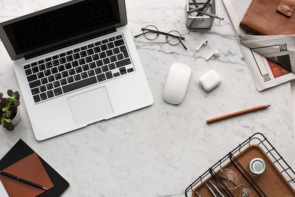 A laptop, computer mouse, a pair of air pods, a pair of reading glasses, and a pencil all sprawled out across a marble desk.