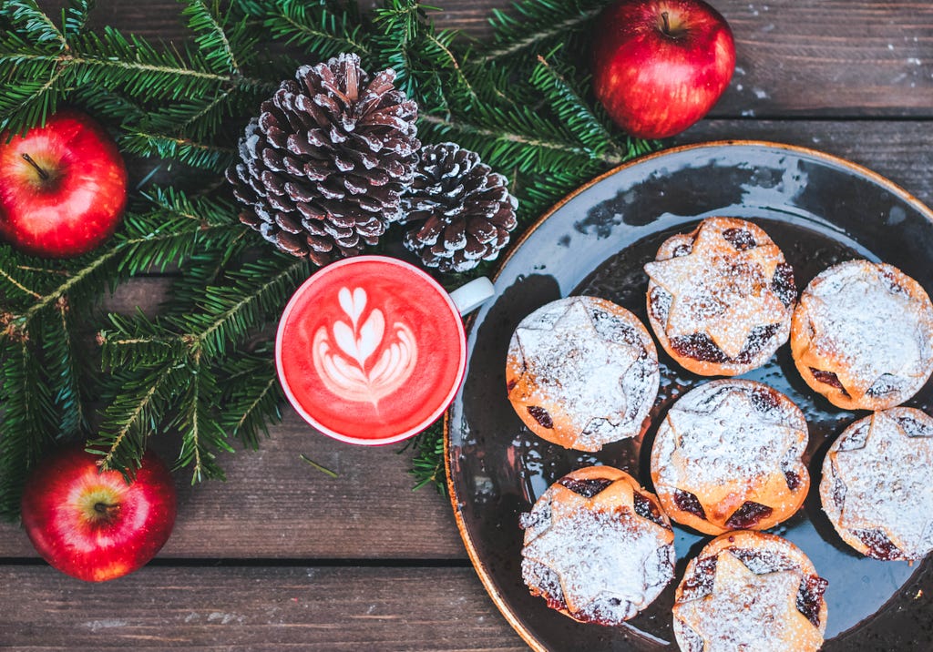 Beautiful sugared tarts rest on a dark-glazed plate on a rustic wood table, next to a red drink in a mug topped with a pretty heart-shaped cream garnish, and a few green bows, pine cones, and red apples.