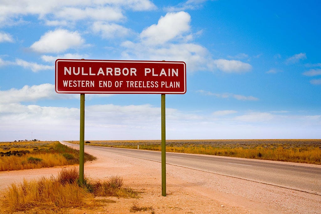 A road sign reading “NULLARBOR PLAIN — WESTERN END OF TREELESS PLAIN” stands beside a paved road under a blue sky with wispy clouds on the Nullarbor Plain.