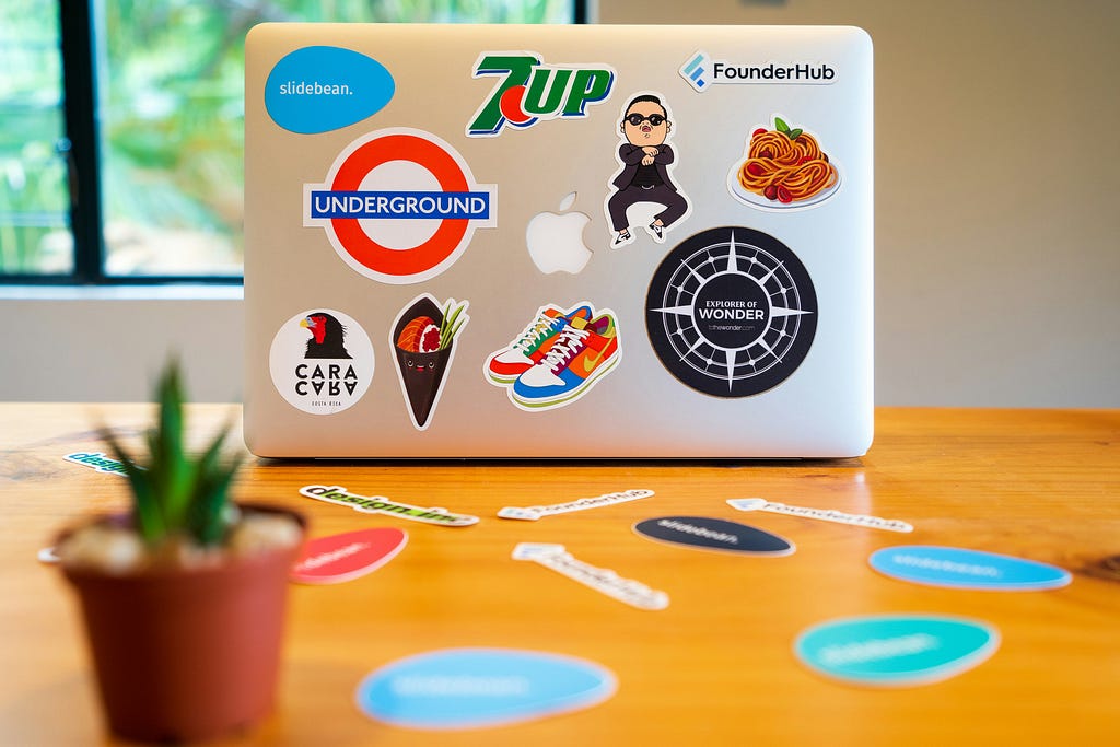 A laptop on a wooden desk covered in various colorful stickers, including logos and fun designs like sneakers and food, with a small potted plant to the left side of the frame and additional stickers scattered on the table