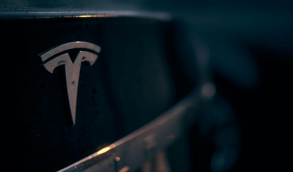 A close up picture of the Tesla logo on the back of a Tesla automotive vehicle.