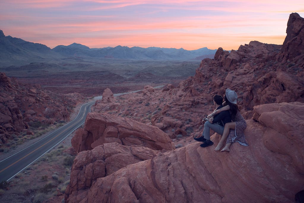 A couple sit together on a red rocky landscape amiring the panoramic view.