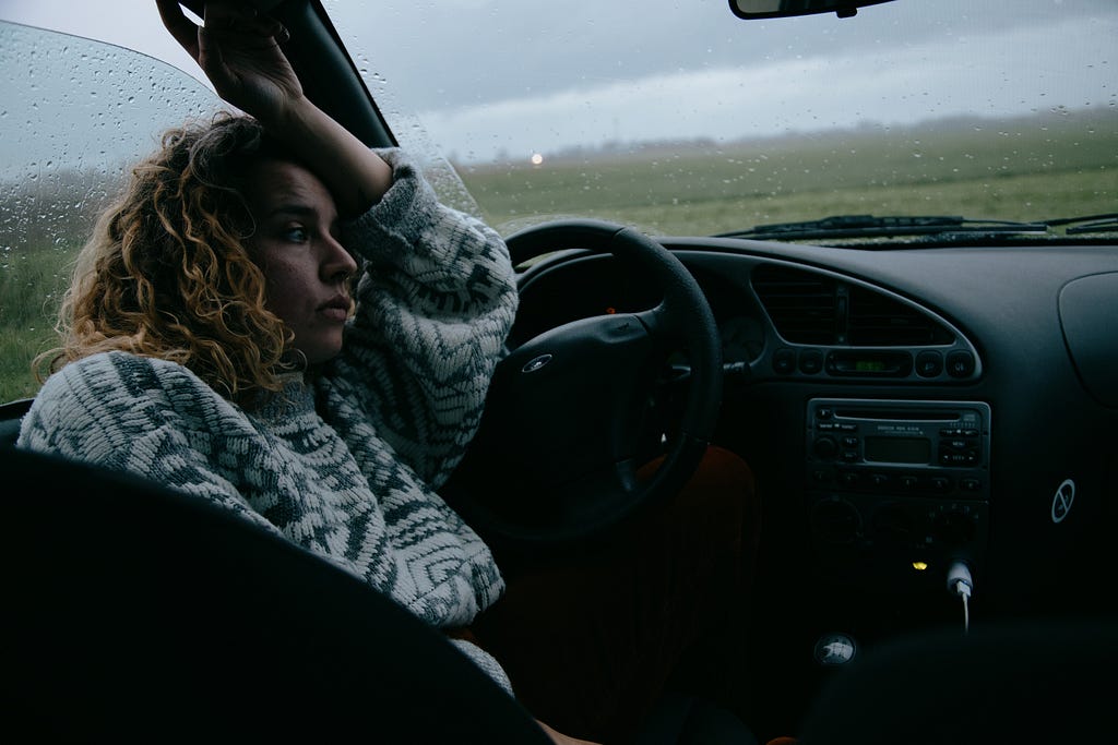 A woman sitting in her car, looking depressed on a dark and rainy day.