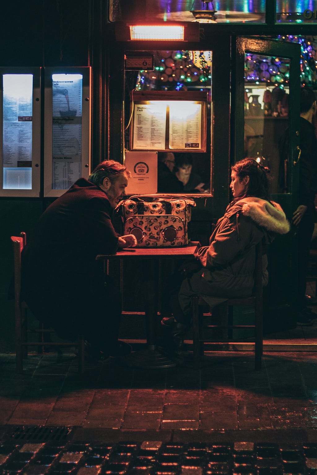 A man and woman sit alone at a table for two, dripping in orange light in an otherwise dark street scene. Inside the bar behind them, crowds celebrate… but these two are serious. They regard each other warily. Something has happened.