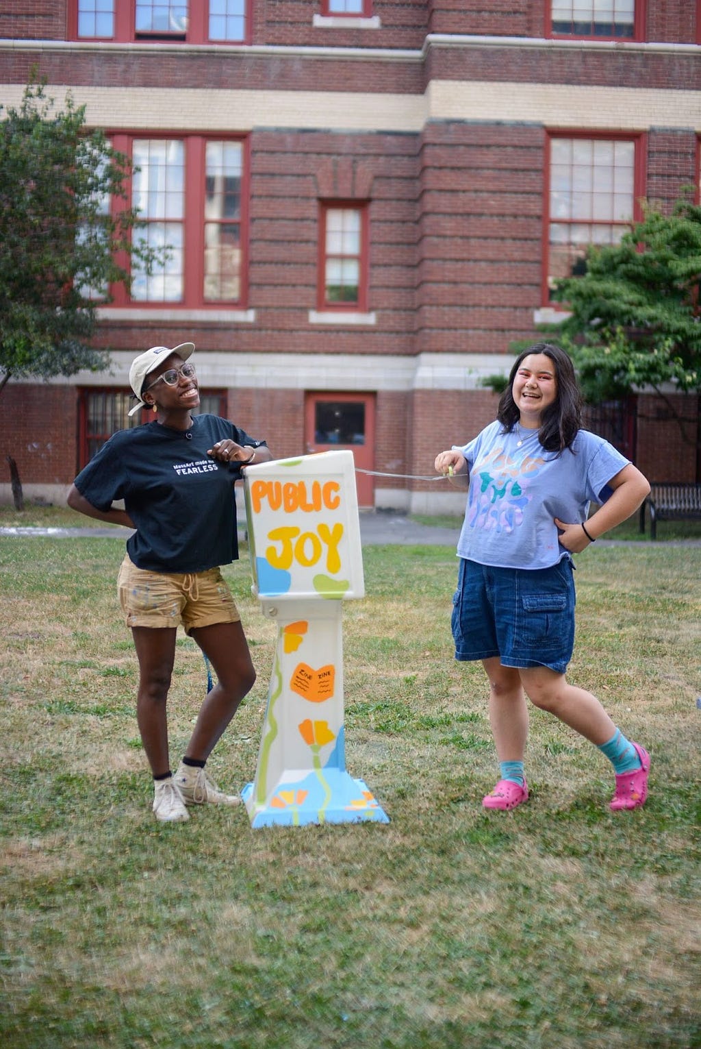 Sabrina and Ava stand with a four-foot-tall box between them on a patch of grass. They are both smiling, and Ava opens the top of the box. The box is painted with bright orange, yellow, green, and blue colors and says the words “Public Joy”.