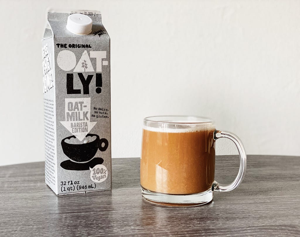 A carton of Oatly Barista centre-left, with a mug of beverage centre-right