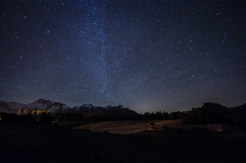 Mountains after dark, silhouetted against a dazzling night sky, with a smattering of glowing stars.