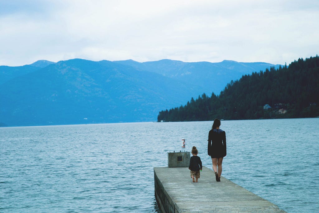 Una madre y su hijo caminan por un muelle, mirando hacia el lago azul, foto enmarcada por montañas