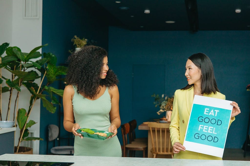 Two women, one is holding a plate with healthy food and the other is holding a poster on which it is written: “eat good, feel good”.