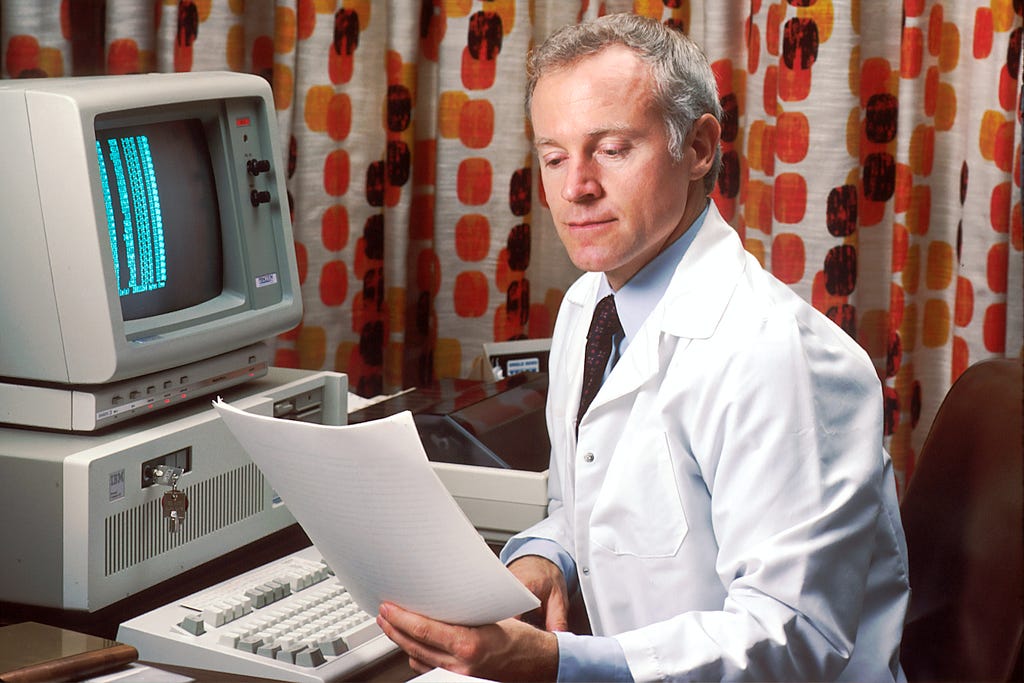 Homem de meia idade trajando jaleco branco observando um relatório de papel, sentado a frente de um computador antigo com tela preta e caracteres brancos.