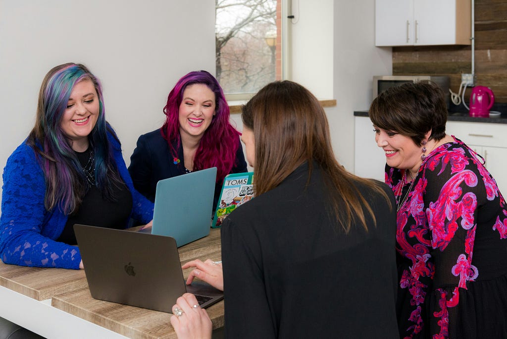 Four women sat round a table smiling, chatting and looking at a laptop screen.
