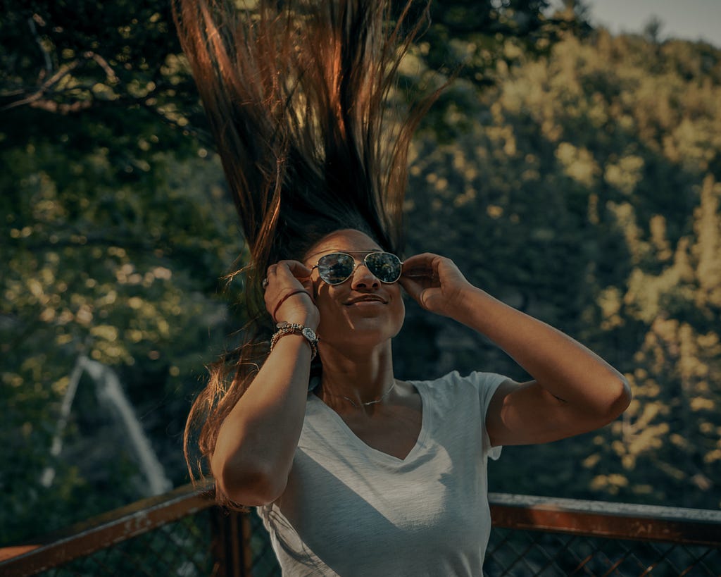 Photo of a woman stands with her back to the corner of a wooden fence with dense green trees in the background. She has flipped her straight hair back so that it looks like it is blowing straight up. She wears a satisfied smirk on her lips while her arms are bent and have set a pair of aviator glasses to her face.
