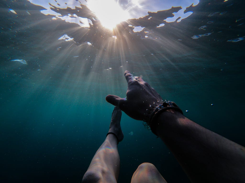 A person, submerged in the ocean, reaches for sunlight streaming through the waves.