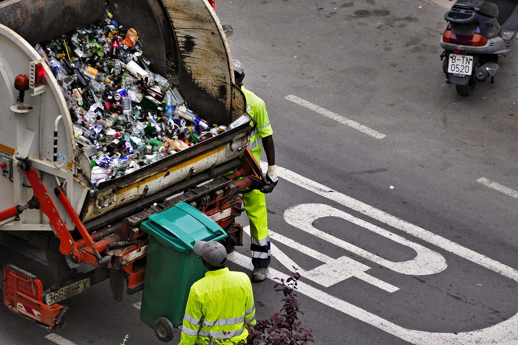 the image is that of the back of a dump truck that is carting away household waste; it is parked in the street and there are two men loading a cart into the back.