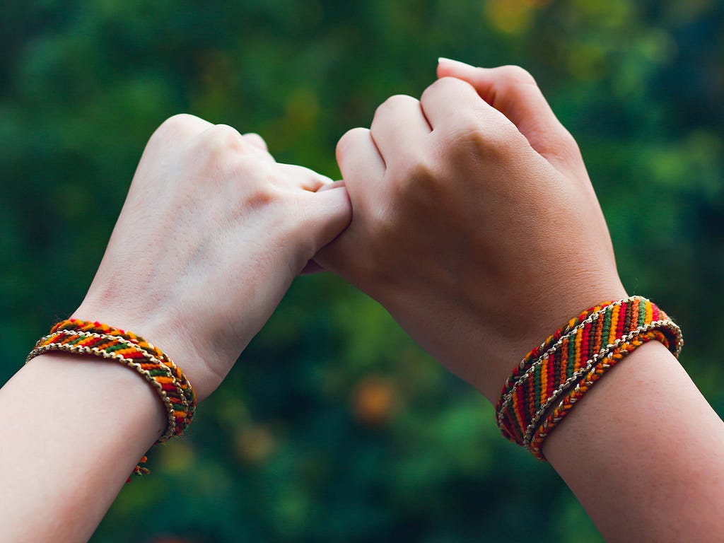 Two hands link with one another’s pinkies. Each hands wrist is wearing a matching friendship bracelet.