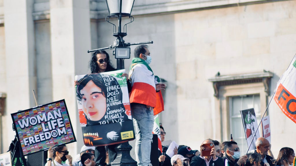 People protest the death of Mahsa Amini. Sign reads: “Woman, Life, Freedom.” Another sign shows a painting of Amini in a hijab and reads: “This piece of fabric does not determine a woman’s right to live.”