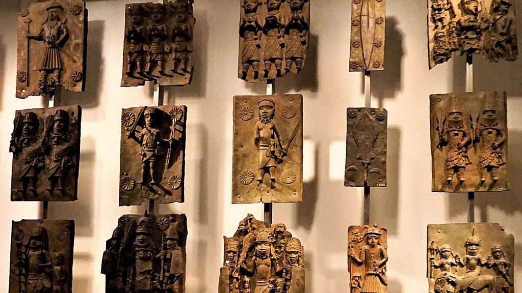 This image displays a collection of Benin bronzes. These intricate metal plaques depict various figures in traditional attire, often engaged in ceremonial or ritualistic activities. The bronzes showcase a high level of craftsmanship with detailed carvings and reliefs, highlighting the rich cultural heritage and artistic achievements of the Benin Kingdom. The plaques are arranged in a grid on a museum wall, with each piece mounted individually for viewing.