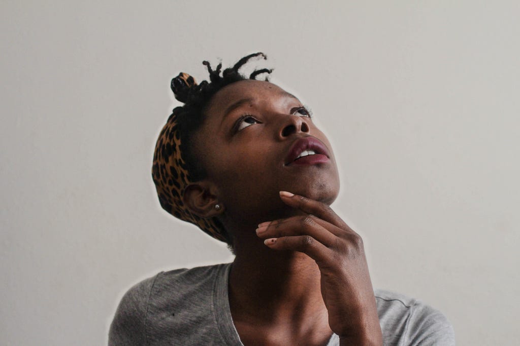 A Black woman with short hair is looking upward and to the left, her left hand under her chin with her chin tilted up. She wears a grey shirt and the background of the photo is also grey.