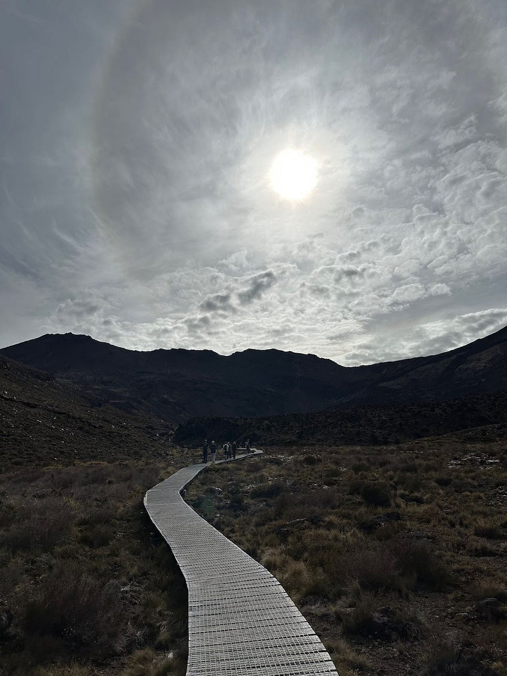 Laid path snaking into distance and hills silhouette with sun in front in sky. Hazy clouds.