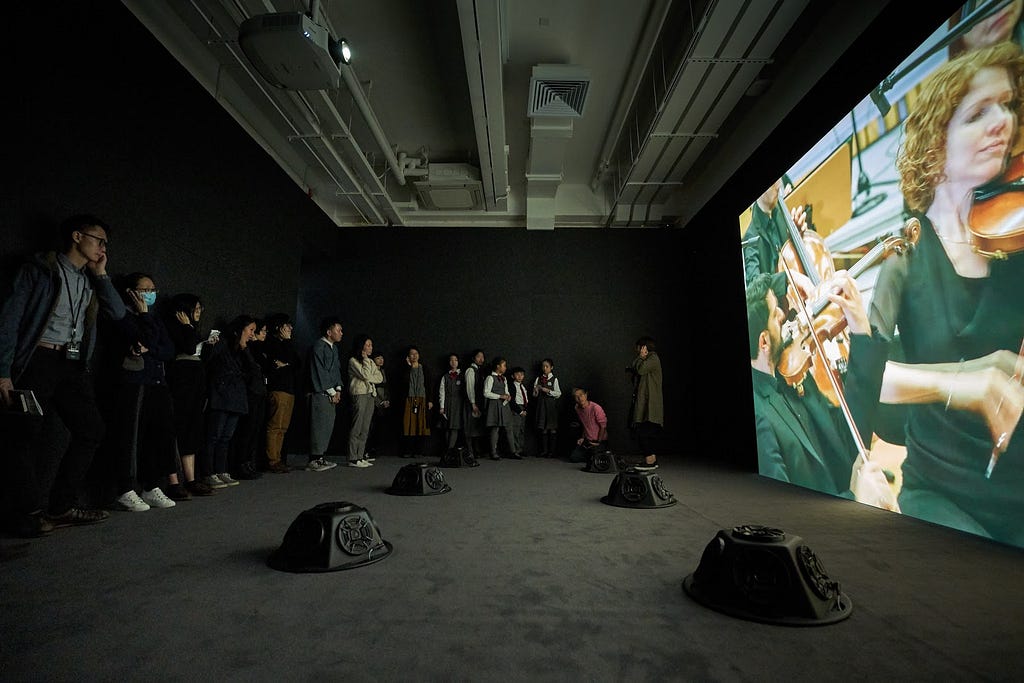 A group of people stand in front of a large screen displaying a video of an orchestra. A young student is speaking.