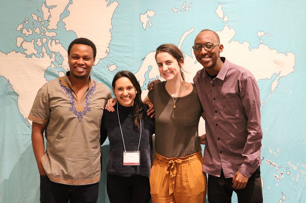 Four smiling people stand together in front of a world map background.