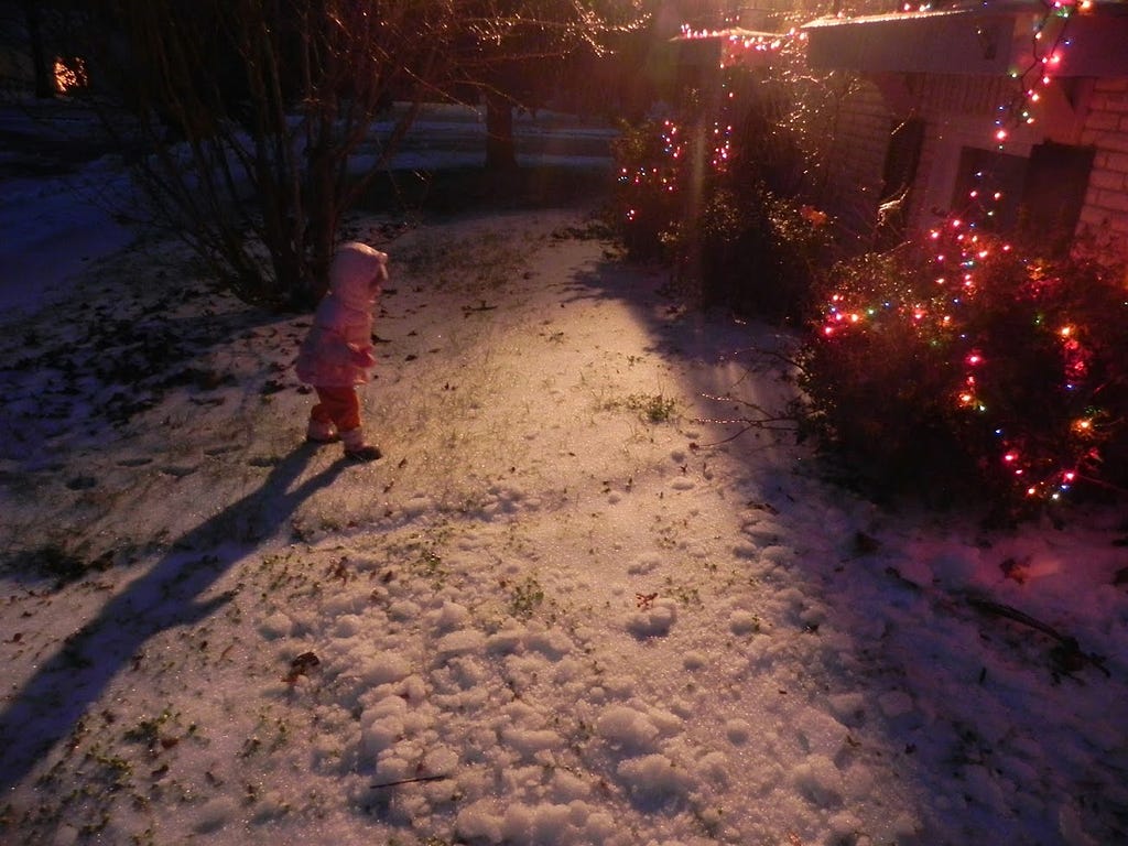 Photo of my daughter, then about 2 years old, in front of our home after a snowy day. Multi-colored string lights strewn over bushes haphazardly.