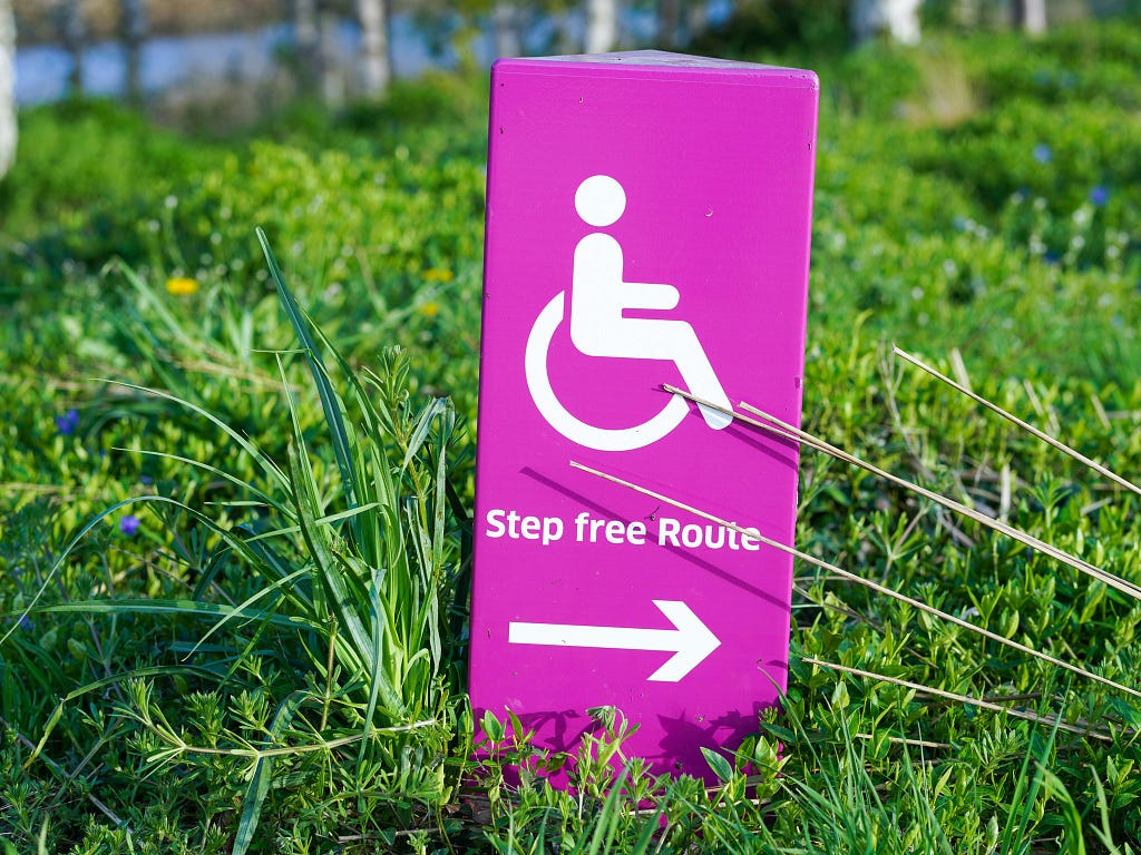Photo of a rectangular lilac object with the disability symbol in white and with a phrase that reads “Step free route”, under this there is a white arrow pointing to the right. The object is on grass.