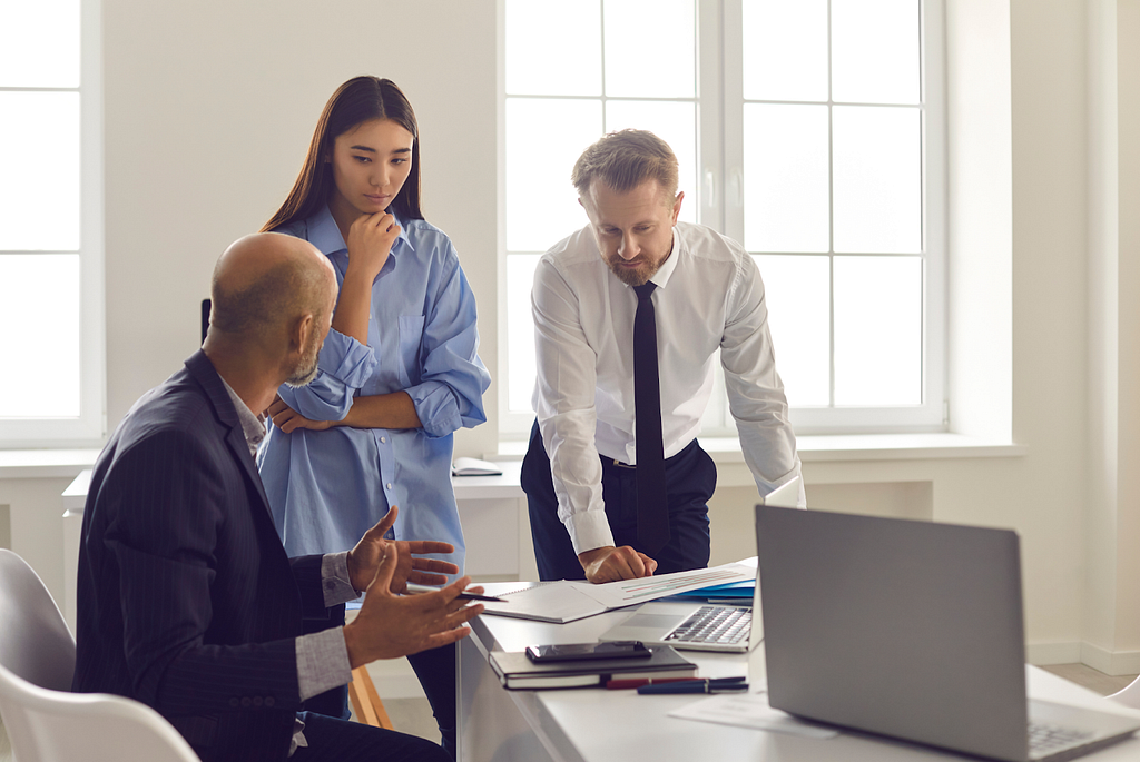 The image shows a manager who is sitting on chair, looking upset towards his 2 employees regarding the reports that they just submitted. It indicates that the team is having an agile roadblock.