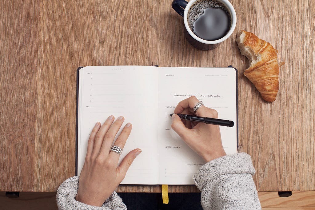A person writing in their notebook with a coffee and croissant nearby