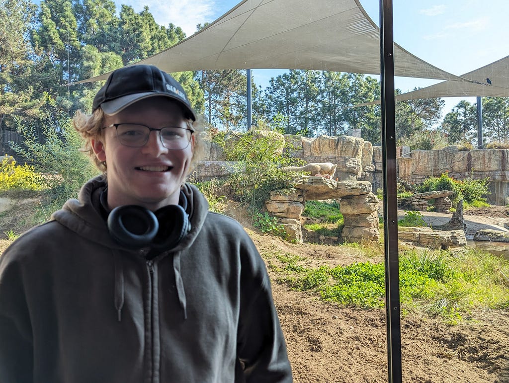 A handsome young man with a ball cap and glasses stands behind the glass of a zoo exhibit. In the background, a polar bear is distantly visible.