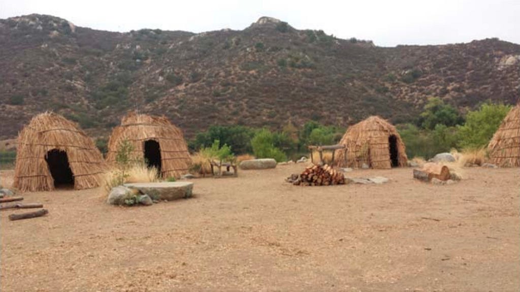 A landscape photograph of a traditional Kumeyaay village of thatched houses with traditional tools, firewood, and structures in a clearing of orange-yellow sandy soil in the foreground, with brownish green chaparral mountain peaks in the background.