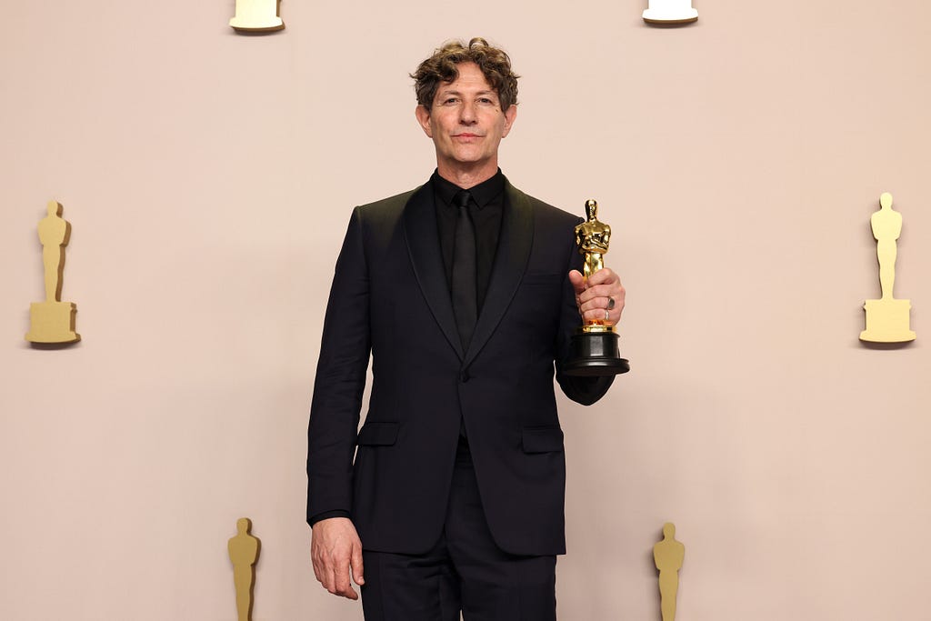 A imagem mostra um Jonathan Glazer dentro do local da celebração do Oscar segurando a sua estatueta do Oscar de Melhor Filme Internacional. Ele está vestindo um blazer e sorrindo.